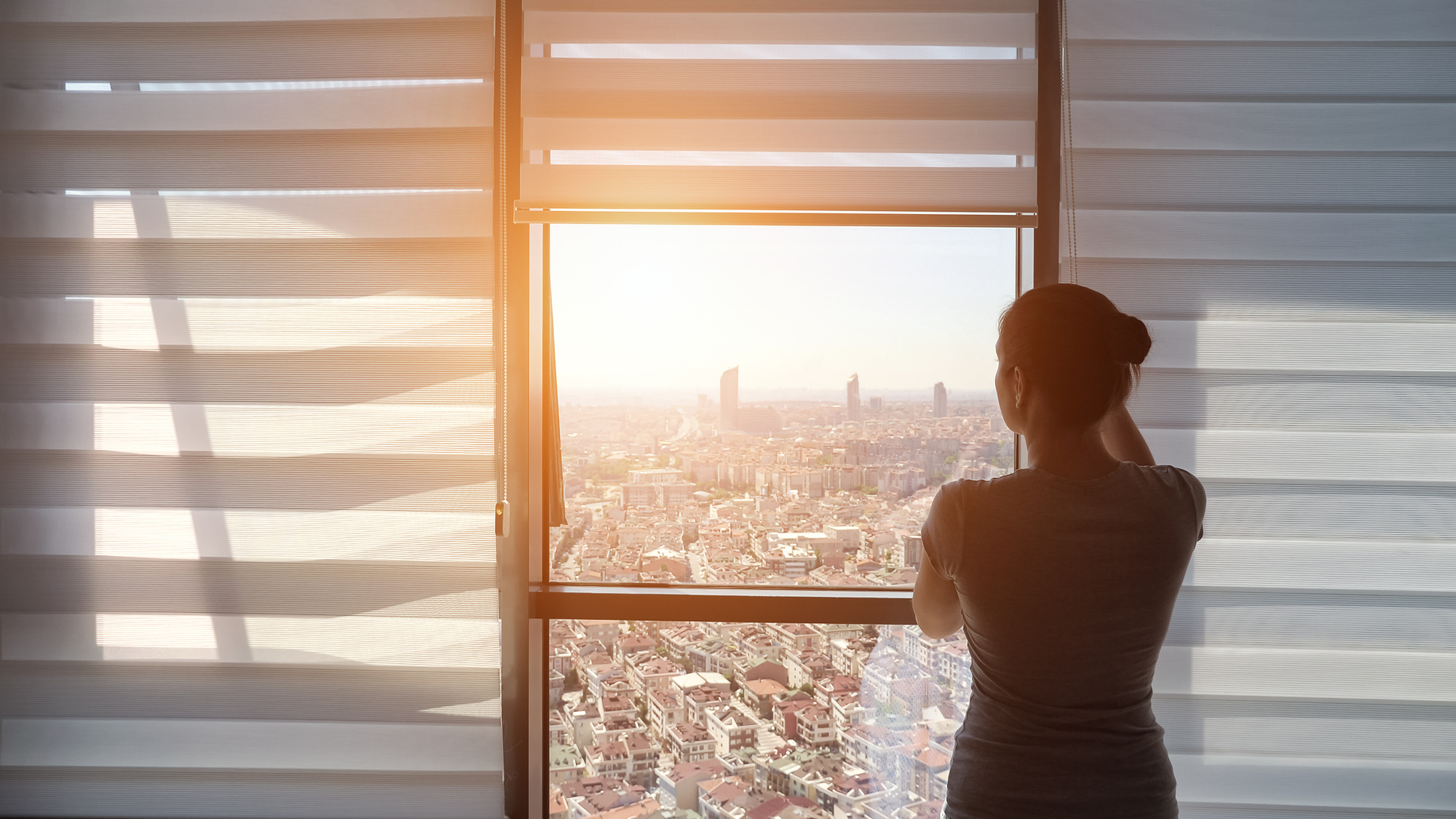 Woman is opening blinds, looking at window with panoramic city view